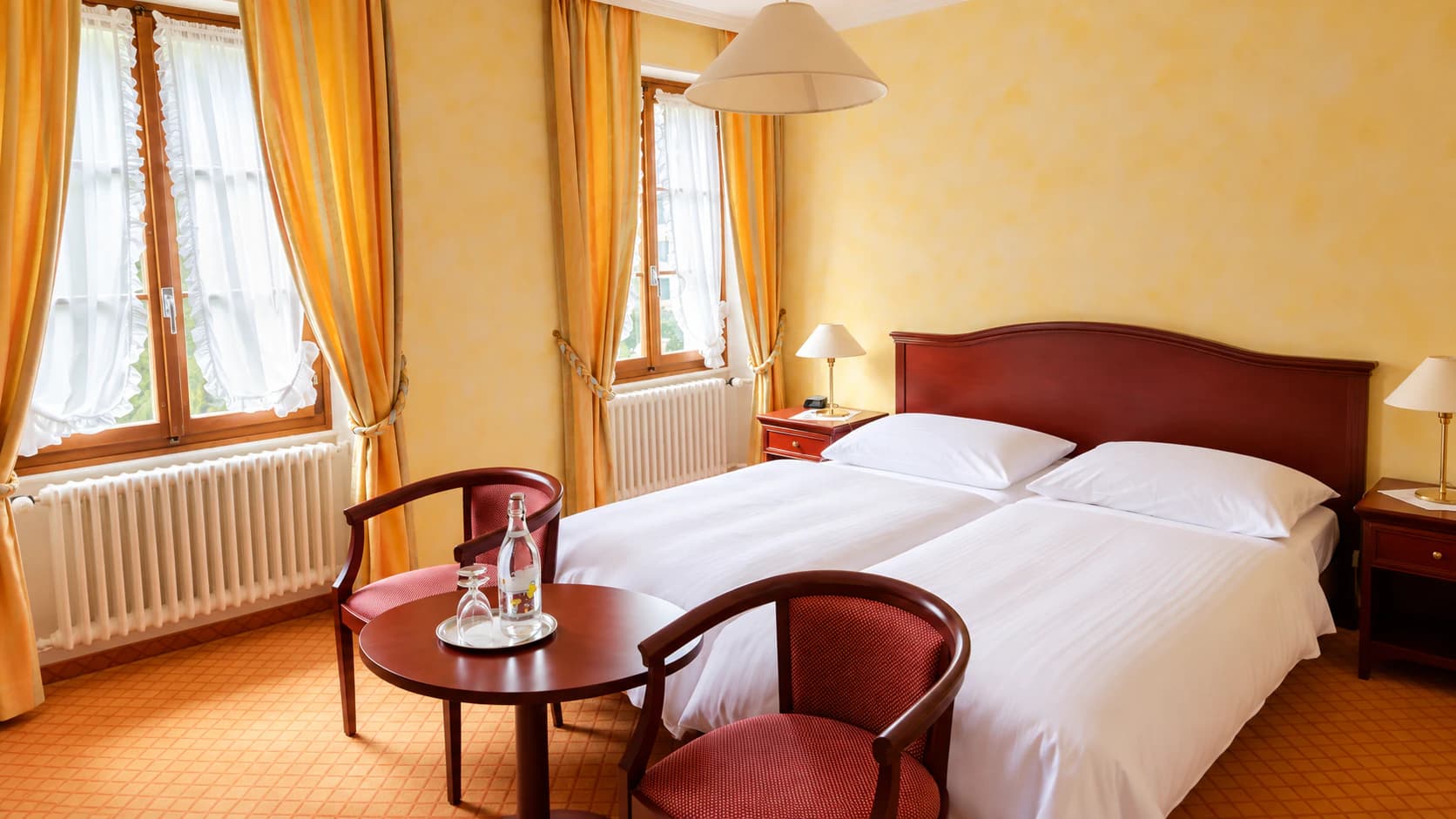Chambre double aux murs jaunes : deux fenêtres, lits jumeaux et coin salon autour d’une table ronde