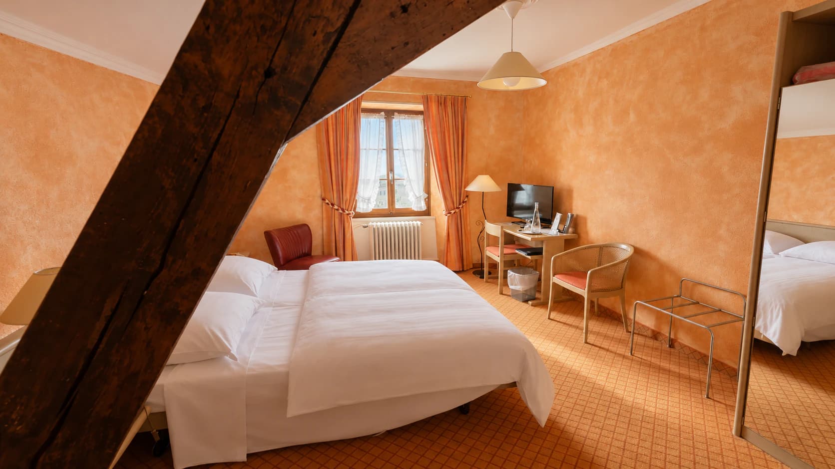 Chambre double aux murs orangés, traversée par une poutre ancienne, avec bureau et télévision
